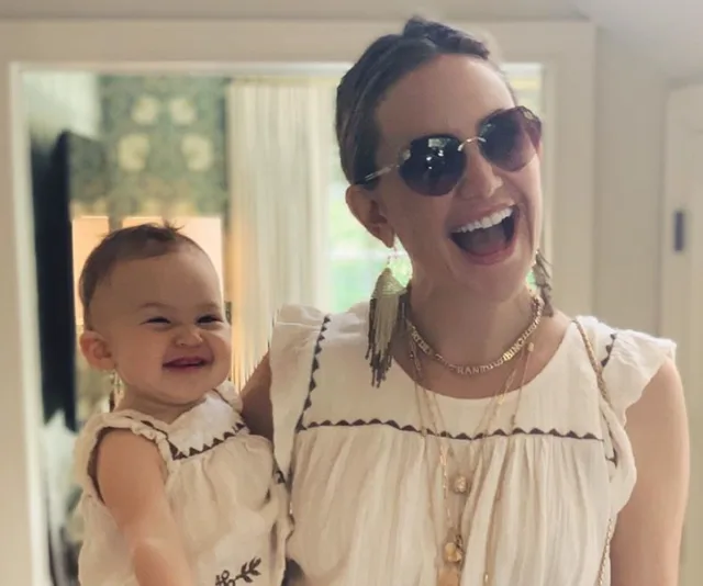 A woman wearing sunglasses, smiling broadly while holding a smiling baby. Both are in matching white outfits.