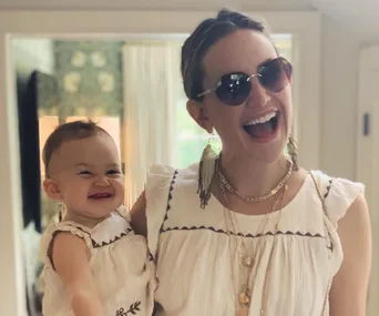 A woman wearing sunglasses, smiling broadly while holding a smiling baby. Both are in matching white outfits.
