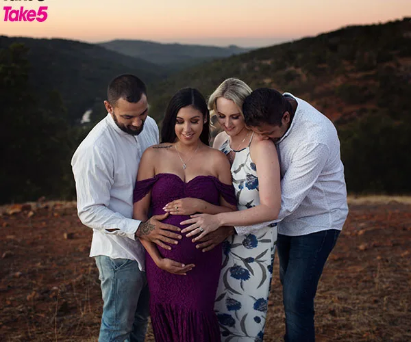 Four adults stand in an outdoor landscape, three gently touching a woman's pregnant belly, with evening hills in the background.