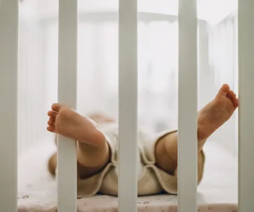 Baby sleeping on back in crib, legs and feet visible through crib bars.