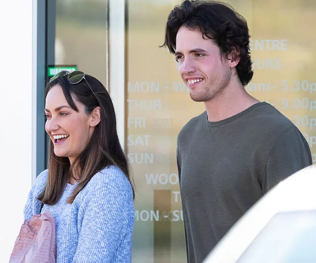 A woman and man smiling outdoors in casual attire near a glass building.