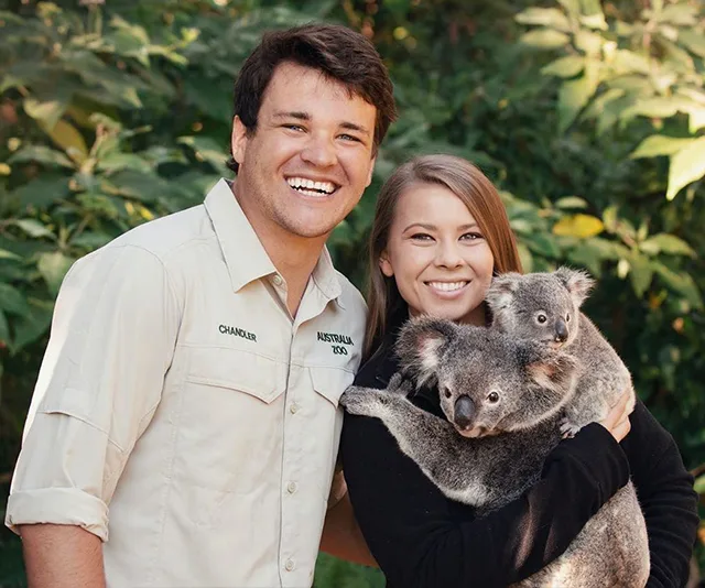 Two people smiling with two koalas in their arms, standing outdoors by leafy trees.