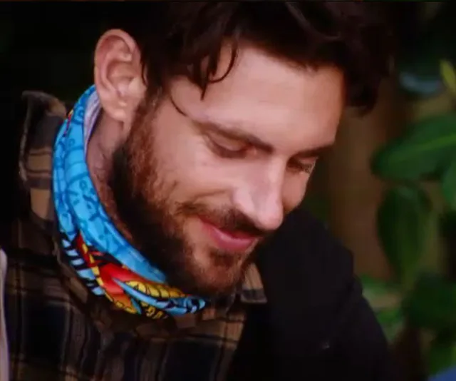 A man with a beard and a colorful neck buff looks downwards, surrounded by greenery.
