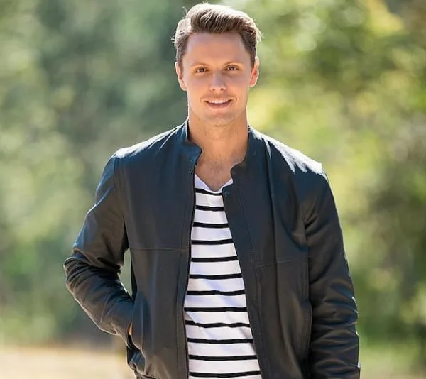 Man in leather jacket and striped shirt smiling outdoors with greenery in background.