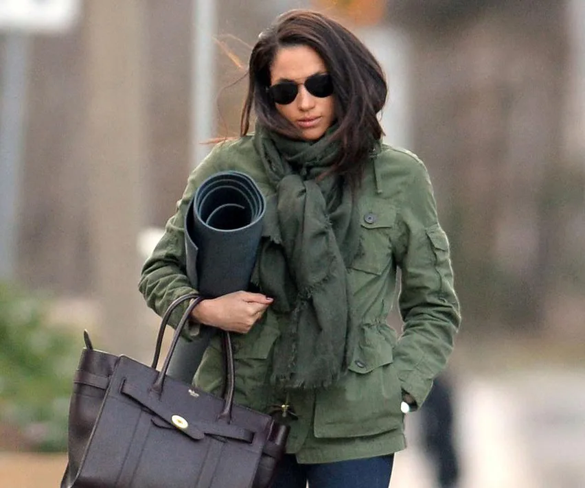 Woman in a green jacket and sunglasses carrying a yoga mat and a handbag outdoors.