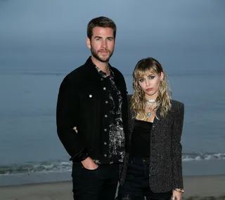 Two people standing on a beach, dressed in stylish outfits, looking towards the camera.