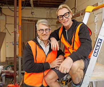 Two individuals wearing glasses and orange safety vests smiling, one sitting on a ladder, in a construction site.