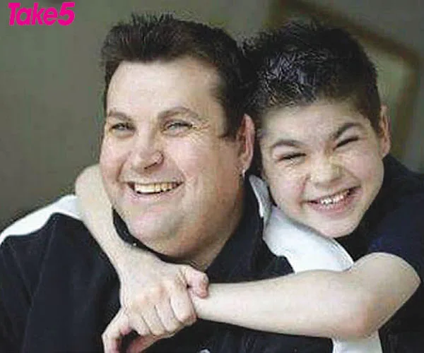 Two smiling individuals embracing, one adult and one child, both with short dark hair. The child is hugging the adult's shoulders.