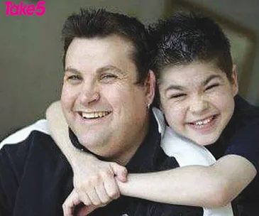 Two smiling individuals embracing, one adult and one child, both with short dark hair. The child is hugging the adult's shoulders.