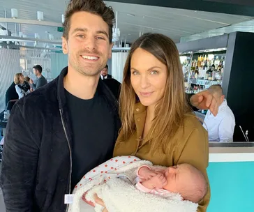 A smiling couple holds a sleeping baby wrapped in a blanket at a modern cafe.