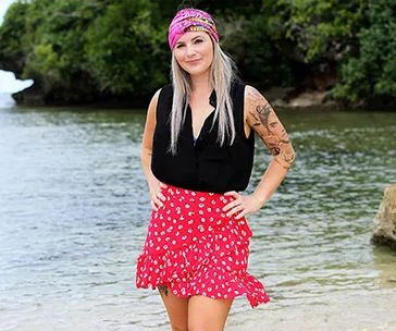 A woman with a pink headband and floral skirt stands on a beach with green foliage in the background.