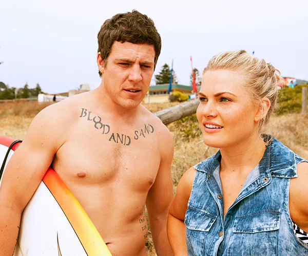 Man and woman standing on a beach, the man holds a surfboard and has a "Blood and Sand" tattoo.