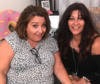 Two women smiling and sitting on a couch, one in a leopard print top and glasses, with a pink donut pillow behind.