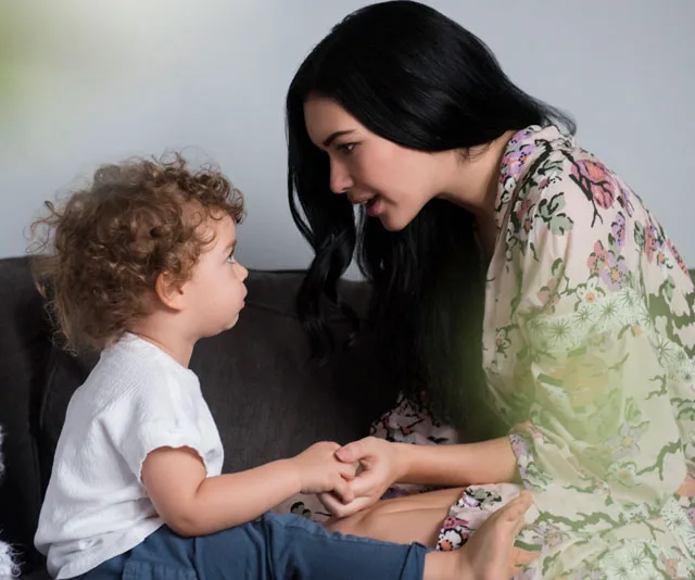 Woman in floral robe holds hands with seated toddler, engaging in a focused conversation on a sofa.
