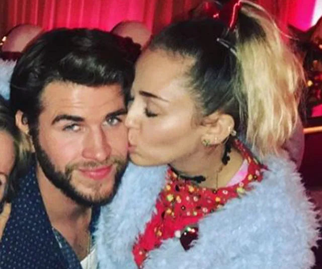 A woman kisses a man on the cheek; both smile gently, seated together at a festive event with pink lighting background.