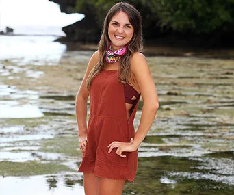A woman standing on a beach, wearing a brown dress and a colorful neck scarf, smiling with hands on hips.