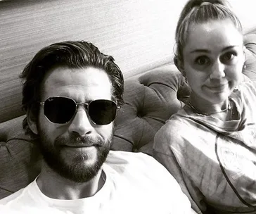 A couple sitting together on a couch; one wearing sunglasses and sporting a beard, the other with a high ponytail, smiling.