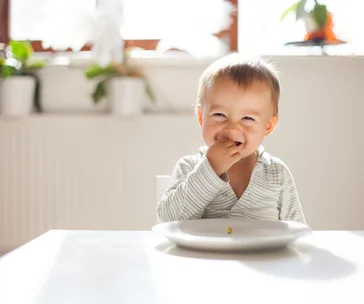 A smiling toddler in a striped outfit sits at a table with a white plate, sunlight and plants in the background.