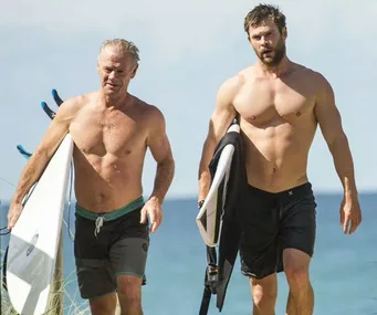 Two shirtless men walking on a beach with surfboards, smiling under clear skies.