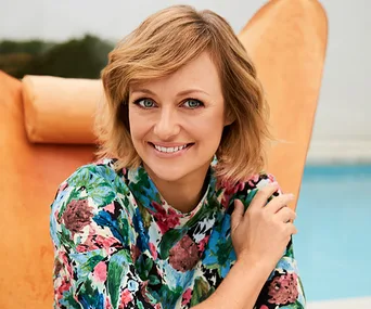 Smiling woman with shoulder-length blonde hair in a floral dress, seated in front of an orange chair by a pool.