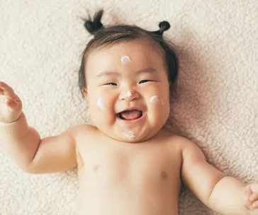 Smiling baby with moisturizer on cheeks and nose, laying on a soft blanket.