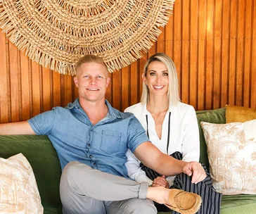A couple sitting on a couch, with a woven decorative piece on the wooden wall behind them.