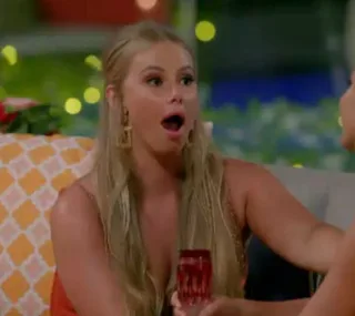 Woman with long blonde hair looks surprised while holding a red glass, sitting on a couch with a colorful cushion.