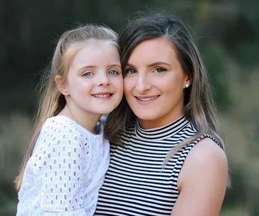 Mother and daughter smiling at the camera, outdoor background.