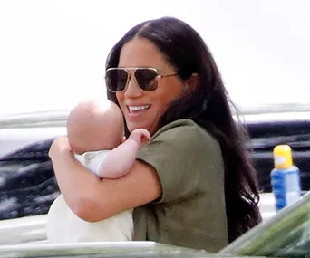 Woman in sunglasses smiling while holding a baby outdoors.