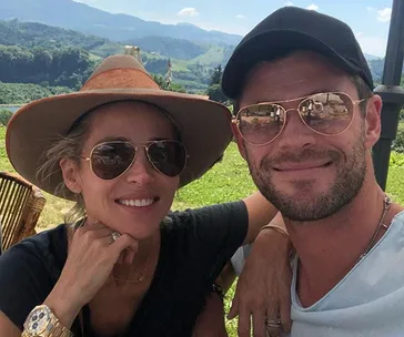 A couple wearing sunglasses and hats smiles for a selfie outdoors with a scenic mountain backdrop.