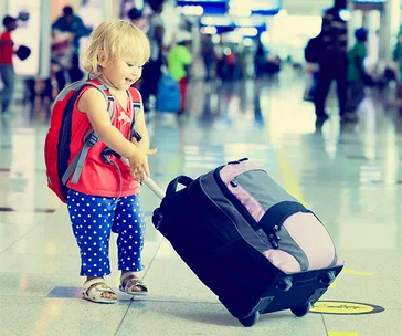 A toddler with a red backpack pulls a suitcase in an airport terminal.