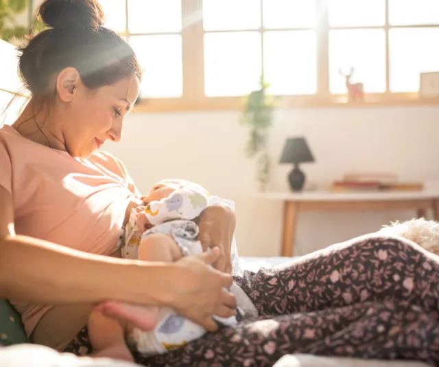 Mother breastfeeding baby in a sunlit room, sitting comfortably on a bed, wearing patterned pants.