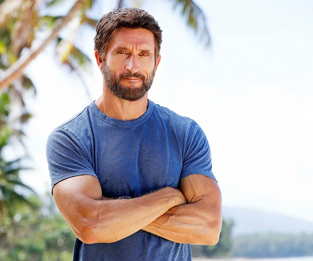 Man in blue shirt stands confidently with crossed arms against a tropical background with palm trees.