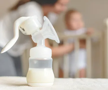 Manual breast pump on a table with a woman and baby blurred in the background.