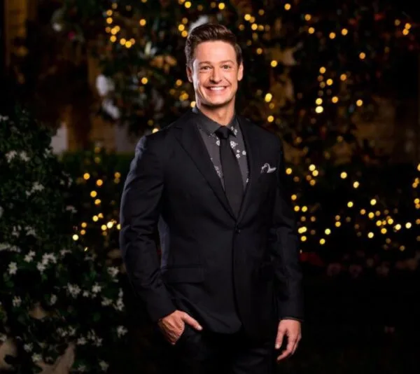 Man in a suit standing outdoors at night, smiling, with decorative lights in the background.