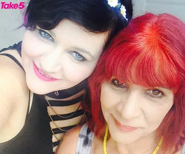 Two women smiling for a selfie; one has black hair with a flower headband, the other has red hair.