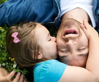 Father and daughter lying on grass, daughter kissing father's cheek, both smiling.