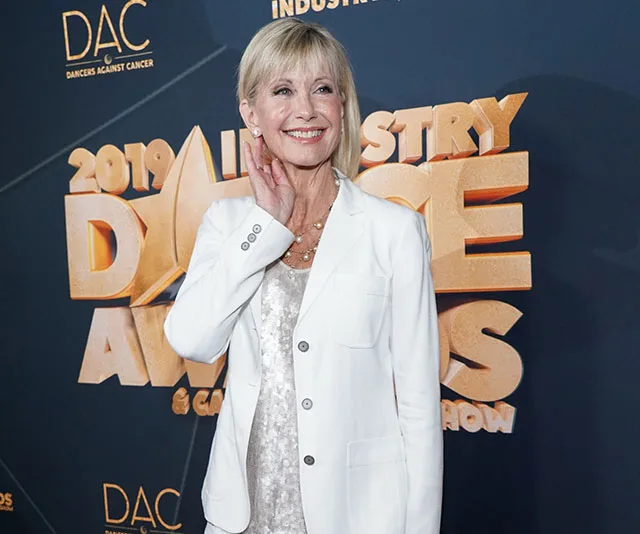 Olivia Newton-John at the 2019 Industry Dance Awards in a white blazer, smiling warmly.