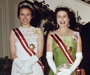 Two women in formal attire and tiaras, smiling, in a ceremonial setting.