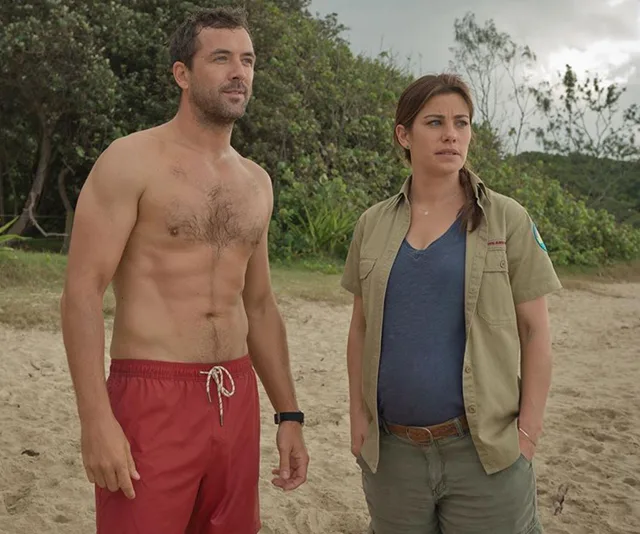 Two people stand on a beach. One is shirtless in red trunks, the other wears a blue shirt and khaki jacket, trees in the background.
