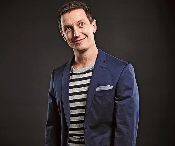 Man in a navy blazer and striped shirt smiling slightly, posed against a dark background.