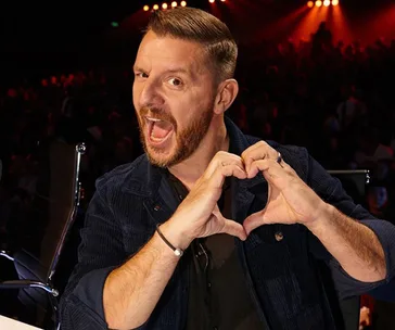 Man making a heart shape with hands, smiling at a talent show, stage lights in the background.