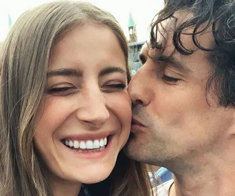 A happy couple is posing for a selfie; one person is kissing the other's cheek while both are smiling broadly.