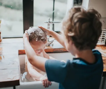 A child gently washes a baby's hair in a sink, with soap suds visible, near a window.