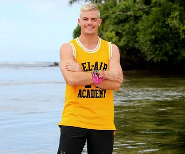 A man in a yellow "Bel-Air Academy" jersey stands on a beach with arms crossed, smiling, backgrounding ocean and greenery.
