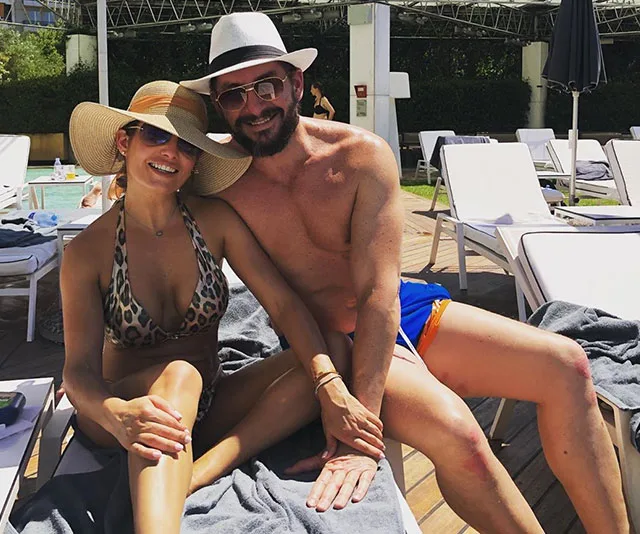 Couple in hats relaxing by the pool on lounge chairs, smiling at the camera.
