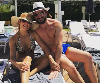 Couple in hats relaxing by the pool on lounge chairs, smiling at the camera.