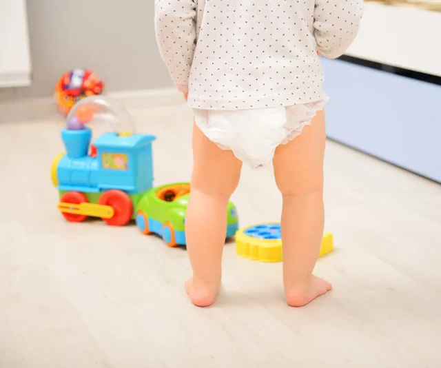 Toddler standing in a diaper, surrounded by colorful toys on a wooden floor.