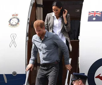 Prince Harry and Meghan Markle descending airplane stairs during a formal arrival, flags and insignia visible.