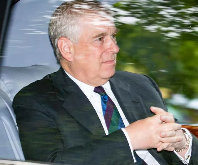 A man in a suit sits in a car with hands clasped, looking to the side.
